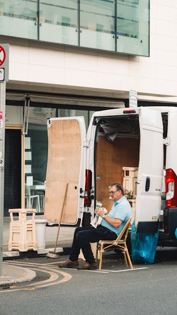 A man with glasses and wearing a light blue shirt is seated on a wooden chair outside a white moving van parked on the street in Limehouse. The van's rear doors are wide open, revealing an interior fi