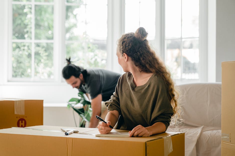 Interior of a home during a house move, showing a woman with long curly hair seated at a table, holding a pen and reviewing documents or a moving checklist for the E14 moving process. In the background, a man with dark hair tied back is lifting or carrying a cardboard box, preparing it for transportation as part of the furniture transport or packing and moving activities. The room is well-lit with natural light coming through large windows, revealing greenery outside. Several packed cardboard boxes, some sealed with tape, are placed on the table and nearby surfaces, indicating ongoing packing tasks related to house removals. A furniture blanket or padding is visible on a sofa, suggesting protection for delicate items during the move. The environment is organized, with moving labels and equipment such as a clipboard and pen being used to coordinate the home relocation. This scene captures the logistical aspects of a professional removals service, as seen in the work of Man with Van Limehouse.