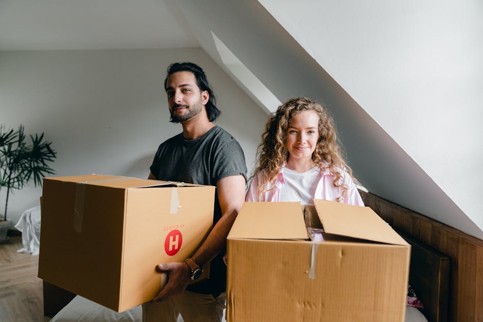 A man and a woman, both smiling gently, stand indoors in a room with white walls and a sloped ceiling, each holding a cardboard box filled with household items. The man, with dark hair and a beard, wears a dark grey t-shirt and carries a large, sealed cardboard box marked with a red 'H'. The woman has long, curly blonde hair and wears a light pink jacket over a white shirt, holding a slightly open cardboard box revealing packed contents. Behind them, a wooden headboard is partially visible, and a potted plant is situated in the corner. The scene is well-lit, suggesting natural daylight, and the furniture and boxes indicate they are in the process of home relocation or packing as part of a furniture transport or moving service. This image exemplifies the logistics involved in house removals, with the individuals engaged in packing or unpacking in preparation for a move, aligned with the services offered by Man with Van Limehouse, as described on their website.