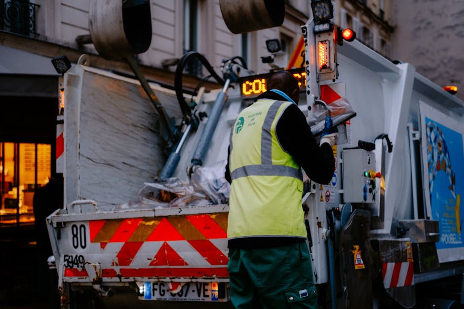 A worker wearing a high-visibility yellow vest and dark clothing is operating a garbage or waste collection vehicle with an open-top container. The worker is facing the vehicle, using a control panel on the side of the truck, which is parked on a street or driveway during dusk or early evening. The container has various materials inside, possibly packaging or waste, with some items wrapped in plastic or cardboard. The vehicle is marked with red and white safety reflective stripes and is equipped with hydraulic or mechanical lifting equipment. The background shows a building with illuminated windows, suggesting a residential or commercial area, aligned with house removals, packing, and waste clearance processes involved in home relocation. Man with Van Limehouse’s service is implied to include efficient waste removal as part of comprehensive house moving support.