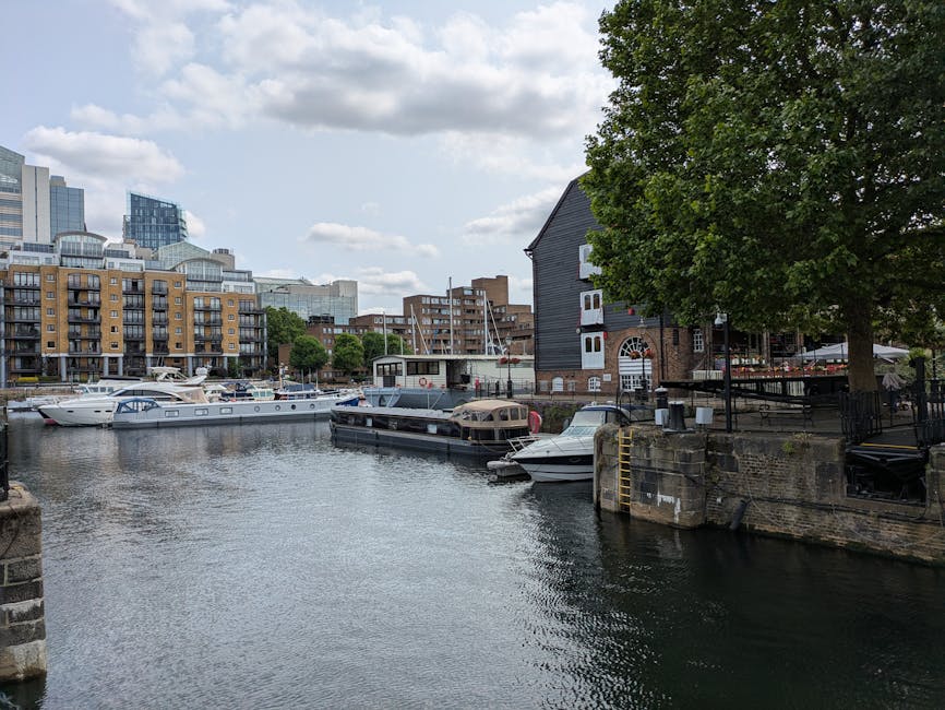 A view of Limehouse Basin featuring calm water with several moored boats and yachts, including a large white motor yacht and smaller vessels, docked along the water's edge. On the right, a dark wooden building with a sloped roof and brick base is situated beside a large, leafy green tree providing shade. The shoreline includes stone and metal railings, with a metal ladder descending into the water. In the background, modern multi-storey buildings with glass and brick facades line the waterfront, indicating an urban environment. The sky is partly cloudy with patches of blue, and the scene captures a quiet moment suitable for home relocation logistics involving boat or furniture transport, facilitated by professional removal services like Man with Van Limehouse.