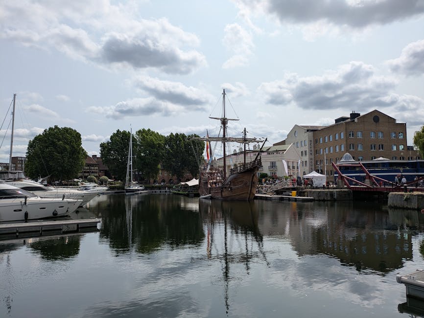 A marina scene during daytime showing several boats docked along the water's edge, including modern motorboats on the left and larger, historically styled sailing ships with wooden hulls and tall masts moored near the shoreline. The sailing ship in the center has rigging and sails lowered, with a few flags visible. Behind the boats, there are trees, a mix of residential and commercial buildings with beige and light-colored facades, and outdoor seating or market stalls set up near the waterfront. The sky is partly cloudy with sunlight breaking through, casting reflections on the calm water. This setting illustrates a location suitable for a house removal or relocation service, with accessible dockside areas and routes for ferrying furniture and belongings, as managed by Man with Van Limehouse.