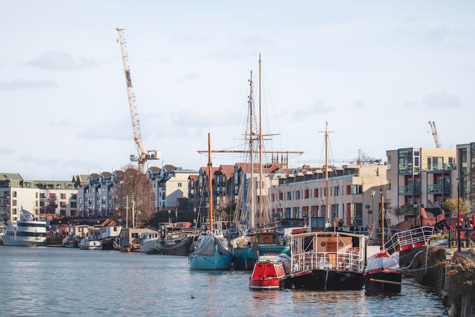 A view of Limehouse Basin featuring calm water with several moored boats and yachts, including a large white motor yacht and smaller vessels, docked along the water's edge. On the right, a dark wooden building with a sloped roof and brick base is situated beside a large, leafy green tree providing shade. The shoreline includes stone and metal railings, with a metal ladder descending into the water. In the background, modern multi-storey buildings with glass and brick facades line the waterfront, indicating an urban environment. The sky is partly cloudy with patches of blue, and the scene captures a quiet moment suitable for home relocation logistics involving boat or furniture transport, facilitated by professional removal services like Man with Van Limehouse.