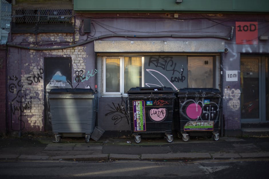 A row of three large wheelie bins positioned on the pavement in front of a weathered building facade. The building's exterior wall features exposed brickwork partially covered with grey plaster and multiple layers of graffiti, including various tags, scribbles, and spray-painted symbols in black, pink, and white. There is a small window with closed blinds situated between the bins, and graffiti continues around it. The bins, used for waste collection, are made of plastic and metal, with the middle one being black and decorated with stickers and graffiti, and the left one in grey without additional markings. The scene is lit by natural daylight, casting soft shadows on the ground. This outdoor setting reflects an urban environment typical for furniture transport or home relocation services, with the bins and building serving as a backdrop for loading or waste disposal during moving processes. The presence of graffiti and urban surroundings indicates the cluttered, real-world context in which care and planning are essential for efficient relocation or removals work, as offered by Man with Van Limehouse.