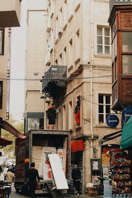 A moving team from Man with Van Limehouse is engaged in a home relocation on a narrow street in Limehouse, with a large moving truck parked adjacent to a multi-storey building. Two movers are visible on an external balcony of the building, both wearing dark clothing and one with an orange cap; one is carrying a piece of furniture or box, while the other appears to be securing or maneuvering items. The truck’s rear doors are open, revealing packing and moving equipment inside, including blankets, straps, and cardboard boxes. Several individuals are on the street, some observing and others assisting, with a variety of shopfronts and local establishments lining the street, including a snack stand with a display of snacks and bottled water. The building’s façade features stonework, multiple windows, and a small decorative balcony, while above, electrical wires run across the scene, indicating typical urban street infrastructure. The environment captures the logistical challenge of navigating a narrow street for furniture transport during a detailed moving process.