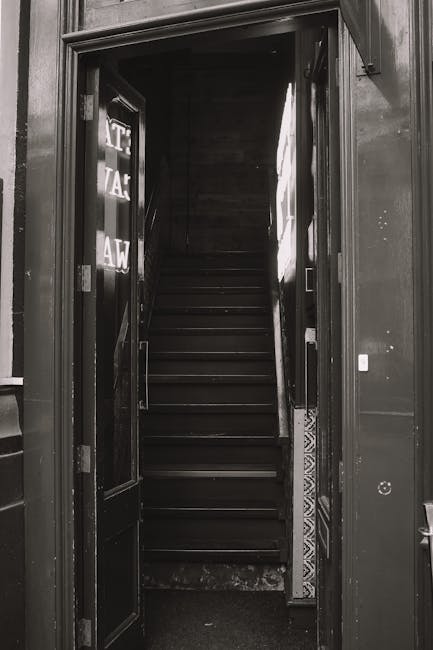A narrow indoor staircase with dark wooden steps and a matching dark wooden handrail, viewed from the bottom looking upwards. The staircase is enclosed by walls, with a small light source illuminating the upper part. To the left of the staircase, a partially visible glass door with the letters 'STA' and 'WA' on it, possibly indicating a staff or service area, is open. The surrounding environment appears to be part of a residential or commercial building entrance, with a small step at the base of the stairs. The image depicts a scene where careful handling is necessary for home relocation or furniture transport through tight spaces, as managed by Man with Van Limehouse, who offers professional removals and moving services.