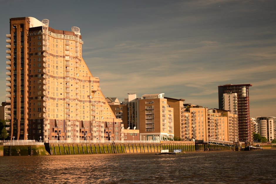 A view of high-rise residential buildings along the riverbank in Limehouse, Tower Hamlets, during daylight with clear weather. The tallest building on the left features a distinctive stepped design with numerous windows and balconies, constructed with reddish-brown brick and concrete. To the right, several other multi-storey apartment blocks are visible, varying in height and architectural style, with some showing external balconies and white window frames. The buildings are positioned along a promenade with a metal railing and a wooden walkway, with a small boat docked on the water. The scene illustrates an urban environment suitable for house removals and furniture transport, with the waterfront setting emphasizing the importance of efficient packing, loading, and logistical planning by providers like Man with Van Limehouse during home relocation processes.