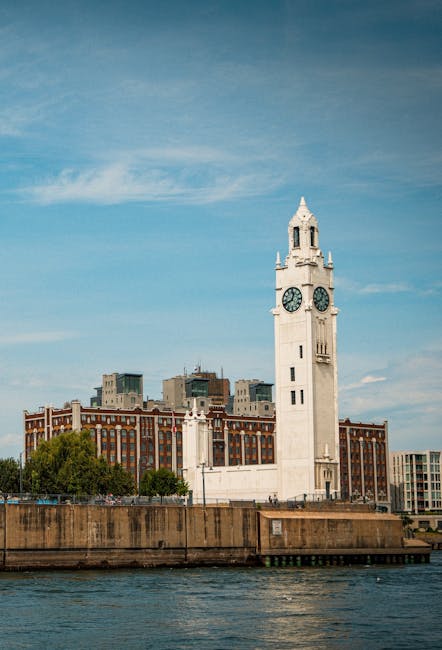 A large white clock tower situated on a brick-lined waterfront with calm water in the foreground. The tower features a prominent clock face near the top, with pointed architectural details and a small spire above. Behind the tower, modern multi-storey buildings with glass and brick facades are visible, set against a blue sky with some wispy clouds. This scene depicts an urban riverside environment, possibly in Limehouse, during daytime, capturing elements associated with house removals and relocating furniture within the area, as managed by Man with Van Limehouse. The setting includes a paved area at the base of the tower, which could be used as a loading or unloading zone during home relocation or moving services.