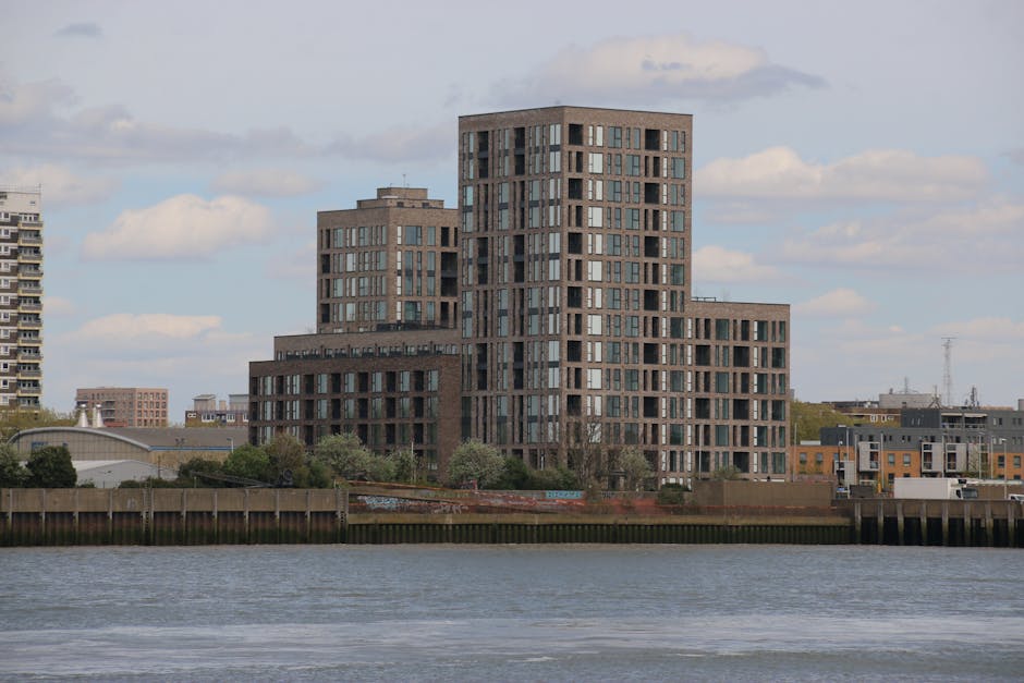 A view of high-rise residential buildings along the riverbank in Limehouse, Tower Hamlets, during daylight with clear weather. The tallest building on the left features a distinctive stepped design with numerous windows and balconies, constructed with reddish-brown brick and concrete. To the right, several other multi-storey apartment blocks are visible, varying in height and architectural style, with some showing external balconies and white window frames. The buildings are positioned along a promenade with a metal railing and a wooden walkway, with a small boat docked on the water. The scene illustrates an urban environment suitable for house removals and furniture transport, with the waterfront setting emphasizing the importance of efficient packing, loading, and logistical planning by providers like Man with Van Limehouse during home relocation processes.