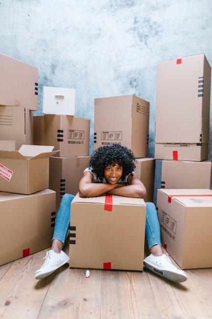 A woman with curly black hair, dressed in a white t-shirt, blue jeans, and white sneakers, is sitting on a wooden floor inside a room, leaning over a medium-sized cardboard box with red packing tape. She is smiling while resting her arms on the box, surrounded by numerous other cardboard boxes of various sizes, some sealed with red or black tape, and others partially open, revealing packing materials or contents. The background features a textured, light blue wall, and the boxes are arranged in a cluttered manner, indicating an ongoing home relocation or packing process. This scene illustrates the logistics of furniture transport, packing, and home removal services provided by Man with Van Limehouse, highlighting the step of organizing and preparing belongings for a house move.