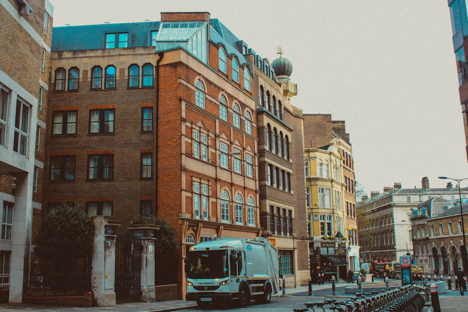 A silver moving van with a sliding side door is parked on a busy urban street beside multi-storey buildings with diverse architectural styles, including red brick and yellow facades. The van is positioned close to a building entrance, with the rear door open, suggesting active loading or unloading. Cardboard boxes and wrapped furniture are visible on the pavement near the van, indicating a home relocation process. The area is illuminated by daylight, with a mix of modern and historic structures lining the street. The scene reflects the typical environment for furniture transport and packing and moving services offered by Man with Van Limehouse, with nearby bicycles and street bollards lining the pavement, emphasizing an organized and accessible setting for residential removals.