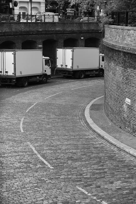 A black-and-white photograph of a curved cobblestone street at the base of a brick retaining wall, with two medium-sized moving trucks parked side by side along the street. The trucks are positioned to facilitate loading or unloading, with the rear doors closed. The street appears to be part of a residential or urban area, with a brick wall on the right and an overpass or bridge above in the background, featuring decorative iron railing and greenery. This scene likely depicts a home relocation or furniture transport process, with the trucks ready for loading or after unloading cargo, possibly associated with professional removals services such as those offered by Man with Van Limehouse.
