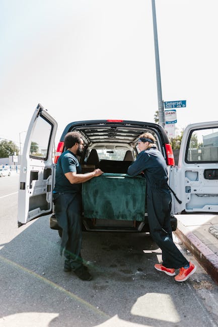 Two professional movers from Man with Van Limehouse are seen loading a large, green, cushioned furniture piece into the open back of a white cargo van on a sunny day. The van is positioned on a paved street with the rear doors swung wide open, revealing the interior space designated for packing and loading furniture. The movers are wearing navy-blue uniforms with safety straps and are carefully lifting the item using proper lifting techniques. In the background, there are street signs, a lamppost, and a row of parked vehicles, indicating a typical urban environment suitable for house removals. The scene captures the logistics of a home relocation, involving careful handling of bulky furniture and transport preparation, reflecting professional furniture transport and packing and moving services offered by [COMPANY_NAME]. The environment is well-lit with natural daylight, emphasizing a smooth loading process necessary for efficient house removals or relocation projects.